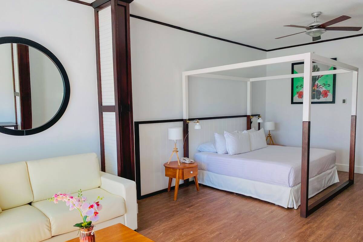 A hotel room with a four-poster bed, a round mirror, a bedside table with a lamp, a small table with a vase of flowers, and a ceiling fan.