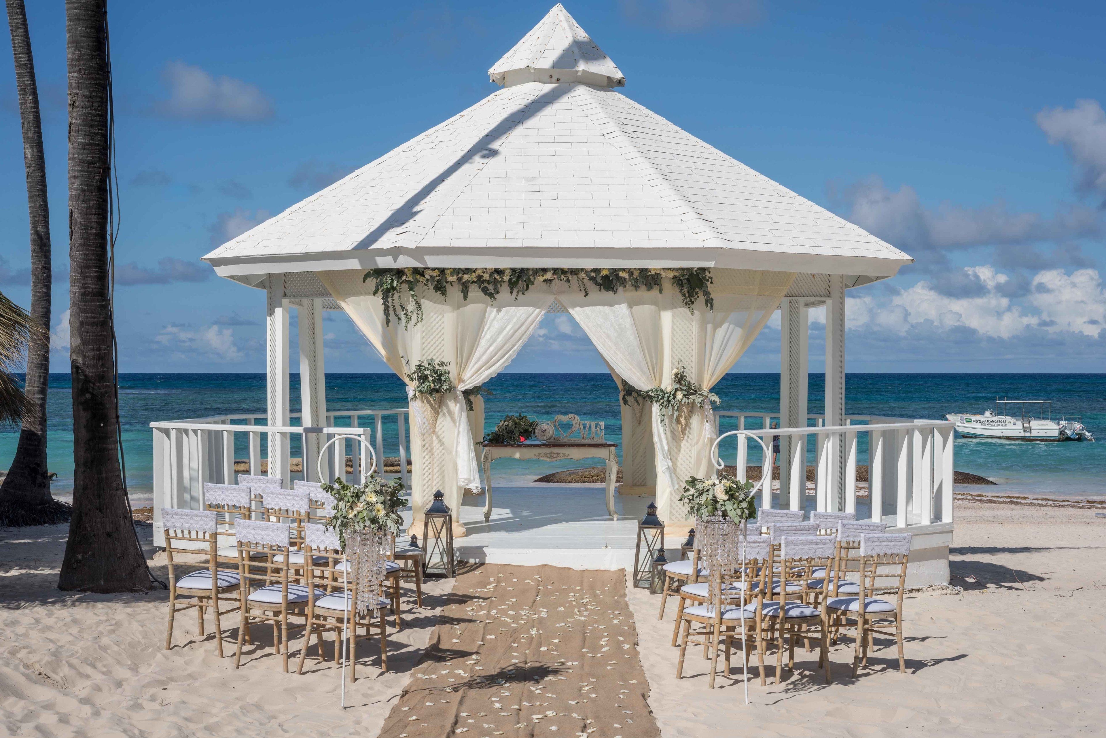 Une cérémonie de mariage organisée sur la plage, avec un gazebo blanc, des chaises et un chemin menant à l’autel.
