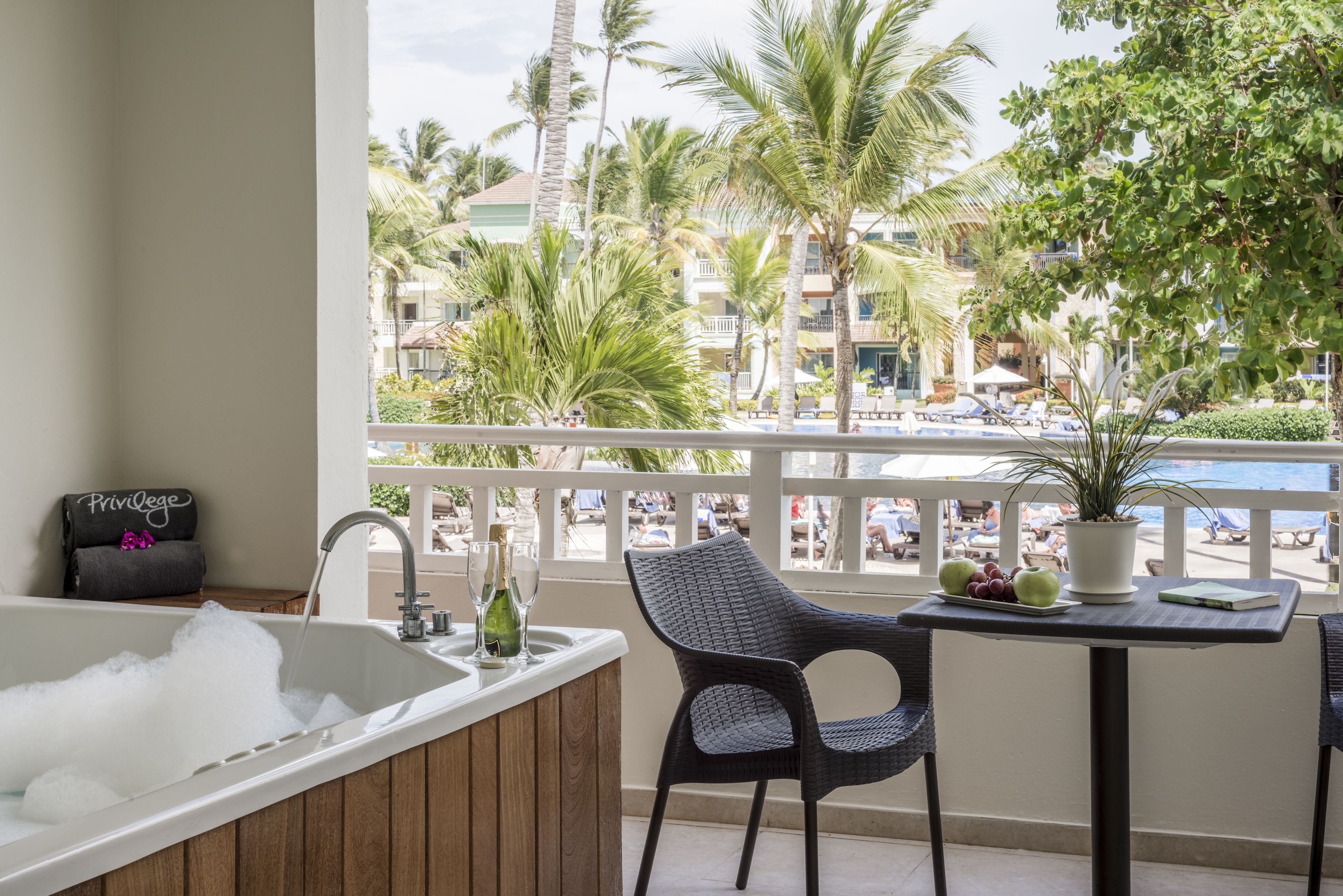 Un balcón con una bañera, una silla, una mesa con planta en maceta y vistas a una piscina y palmeras.
