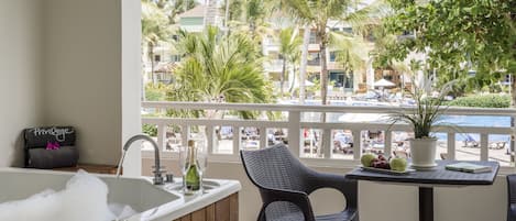 Un balcón con una bañera, una silla, una mesa con planta en maceta y vistas a una piscina y palmeras.
