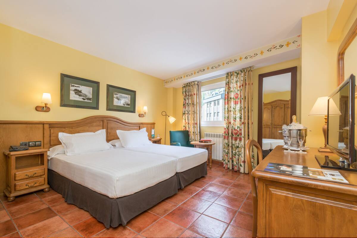 Habitación de hotel con una cama doble, dos mesitas de noche, una pequeña mesa con lámpara, una silla, un escritorio con espejo y una ventana con cortinas.