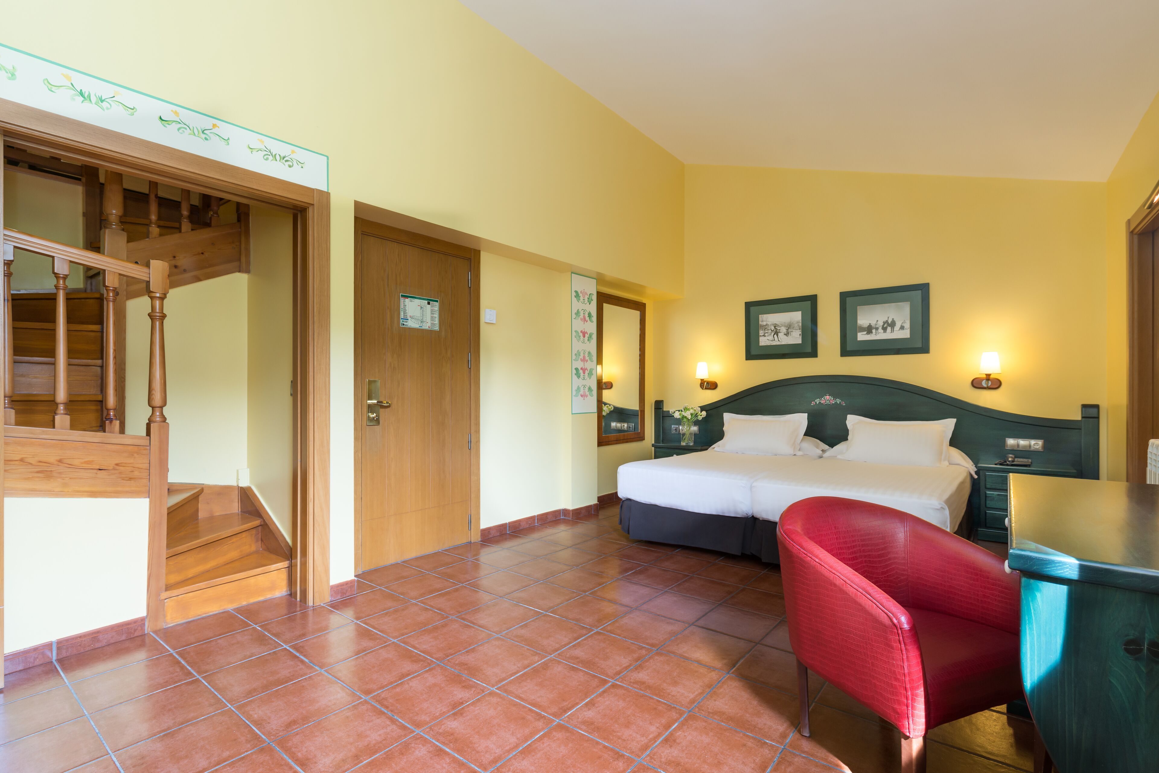 A hotel room with a wooden staircase, a bed with a dark headboard, a red chair, and a desk.