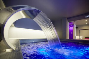 Una piscina interior moderna con una original estructura curva de agua y una zona de estar con una pared iluminada en morado.