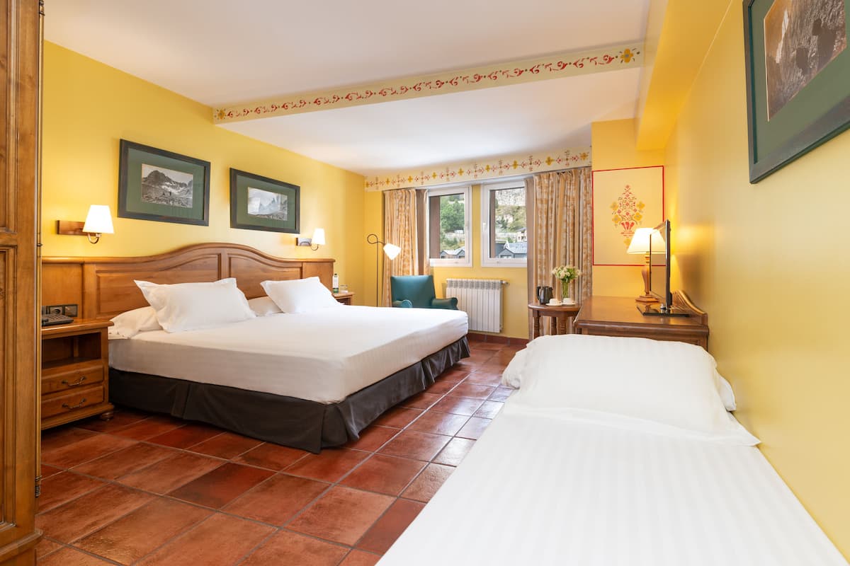 Habitación de hotel con dos camas, una mesita de noche de madera, una silla, un ventanal con cortinas y cuadros enmarcados en la pared.