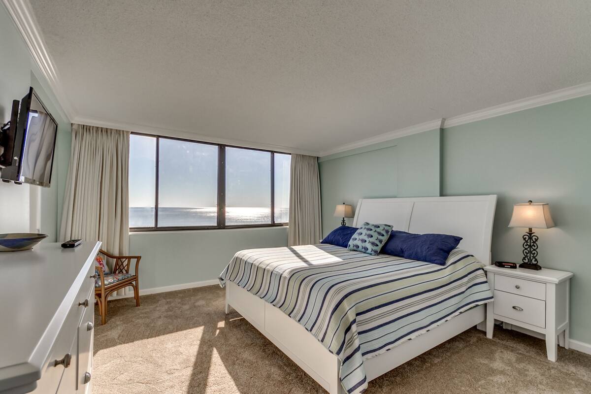 A bedroom with a bed, nightstands, a TV, a chair, and a view of the ocean.