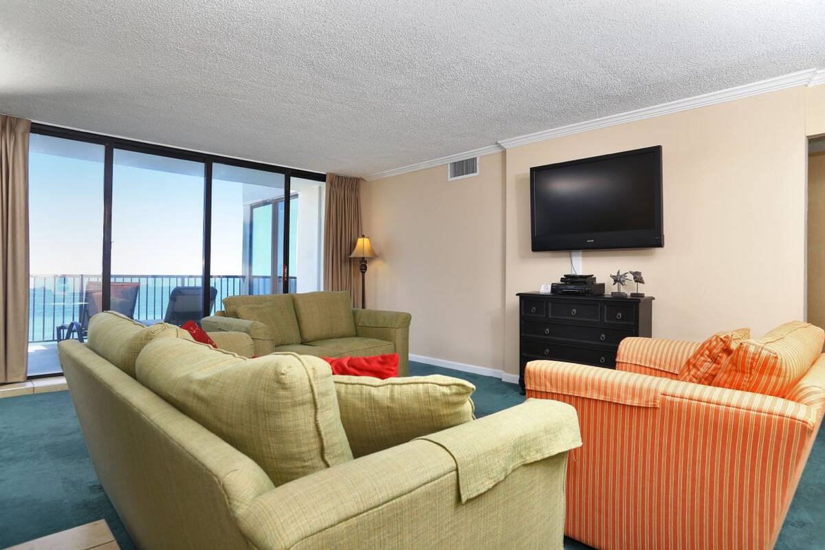 A hotel room with a large window, a flat-screen TV, a sofa, a chair, and a small table.