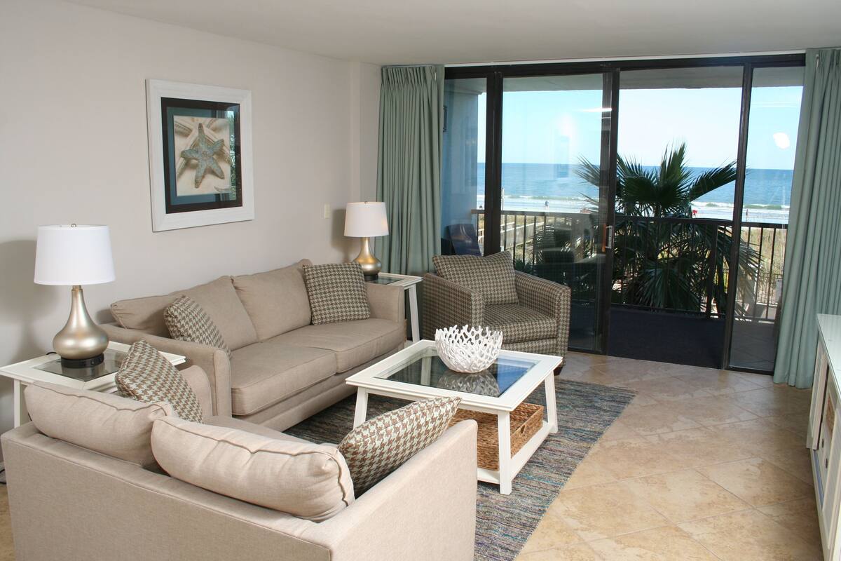 A living room with a beige sofa, a glass coffee table, and a view of the ocean through large sliding doors.
