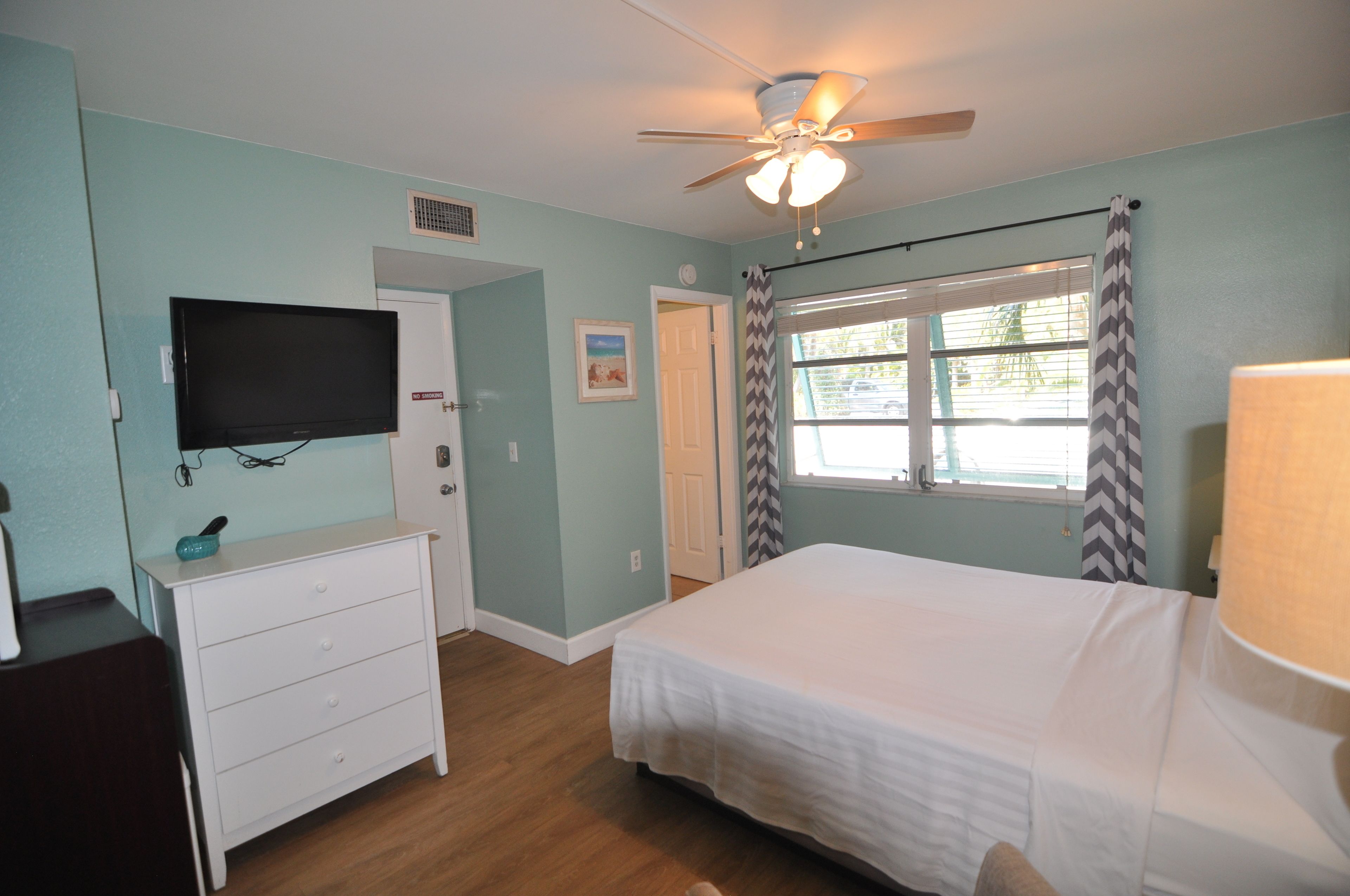 A bedroom with a bed, a ceiling fan, a television, a dresser, and a window with curtains.