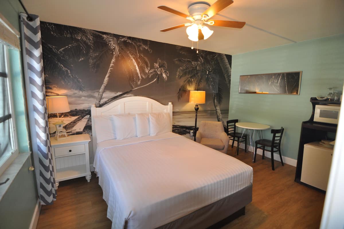 A bedroom with a bed, a ceiling fan, a nightstand, a lamp, a small table with chairs, and a TV.