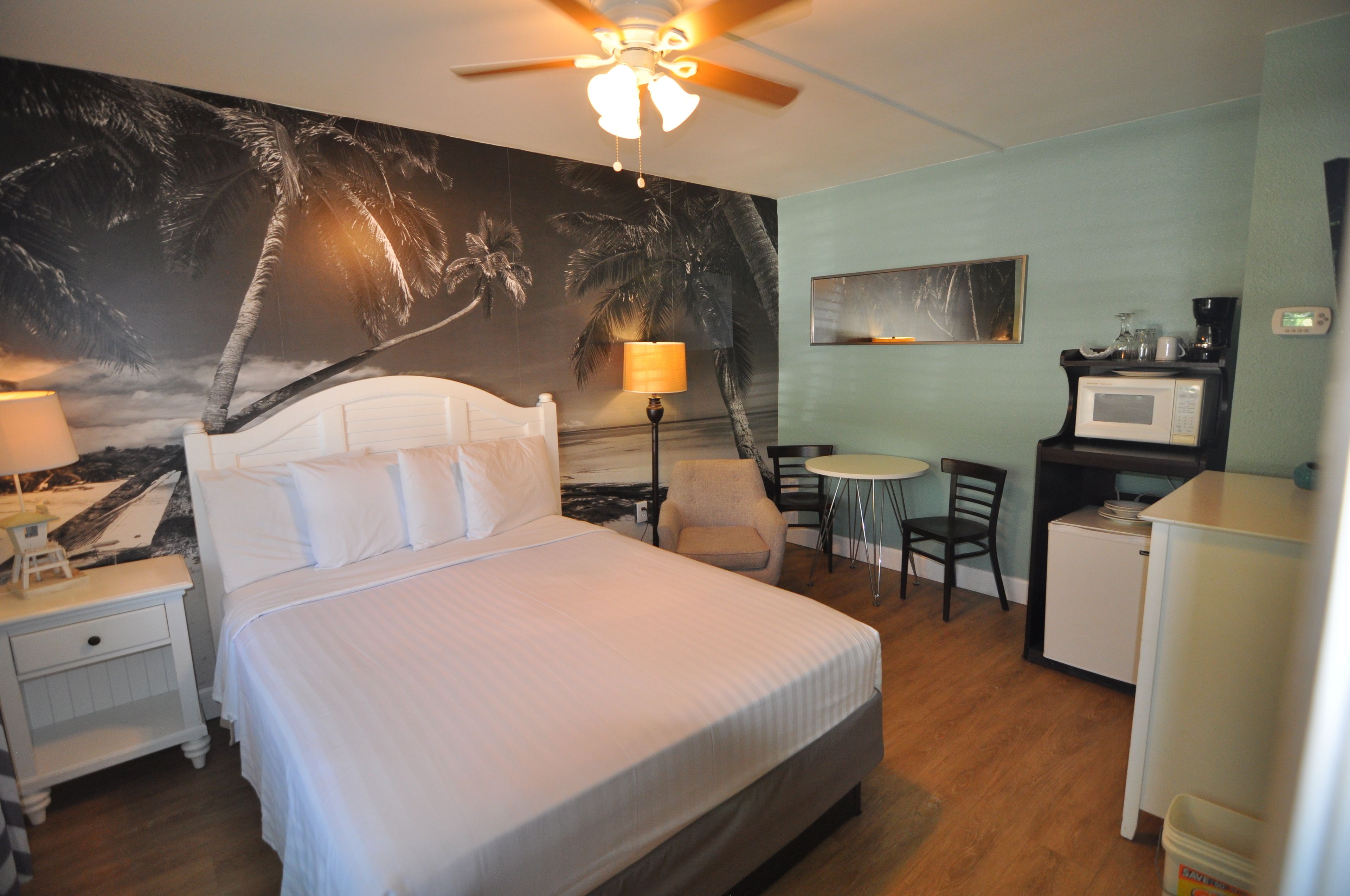 A hotel room with a bed, a desk, a chair, a small refrigerator, a lamp, and a ceiling fan.