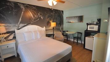 A hotel room with a bed, a desk, a chair, a small refrigerator, a lamp, and a ceiling fan.