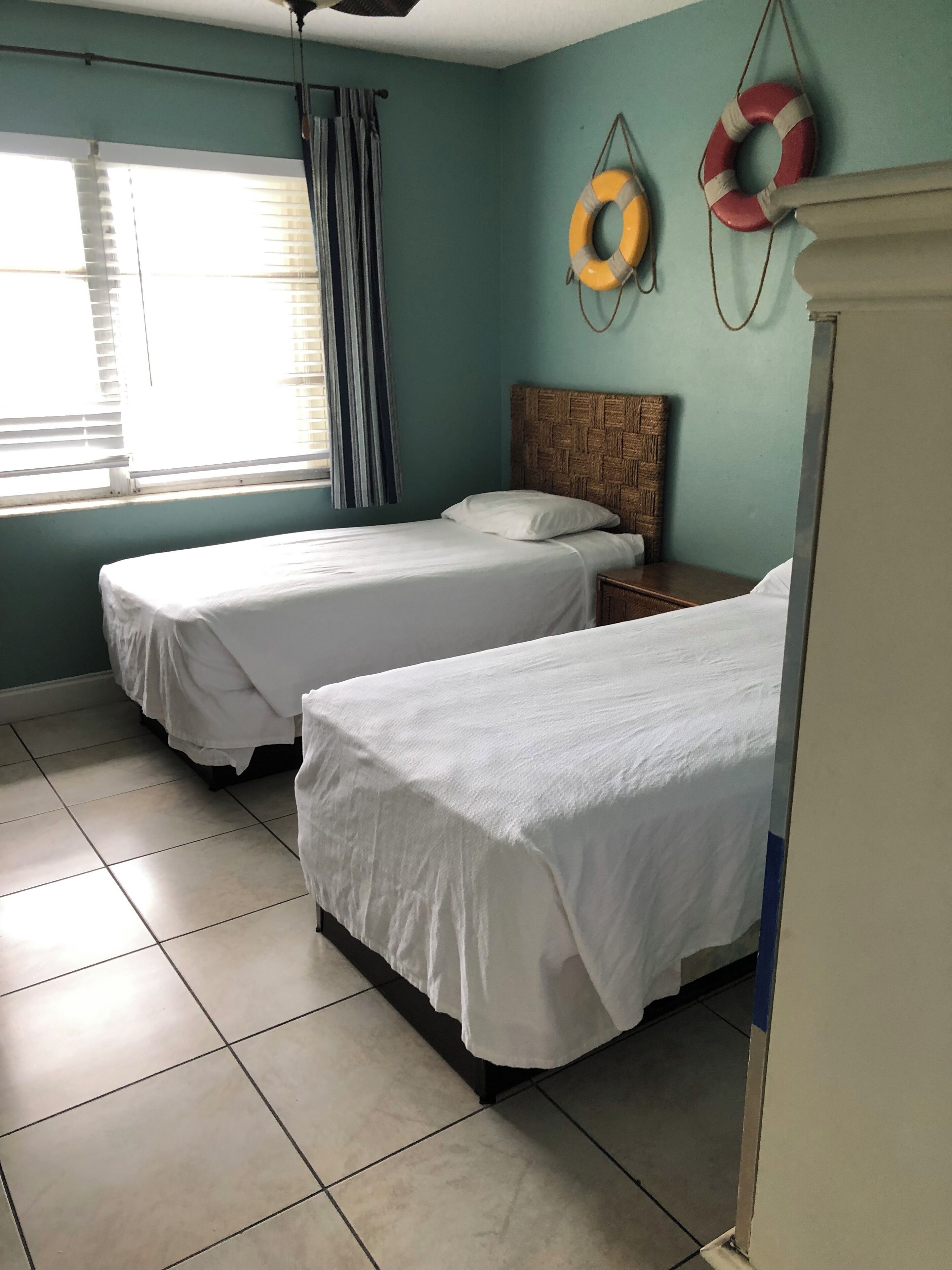 A room with two beds, a window with blinds, a ceiling fan, and a life ring on the wall.