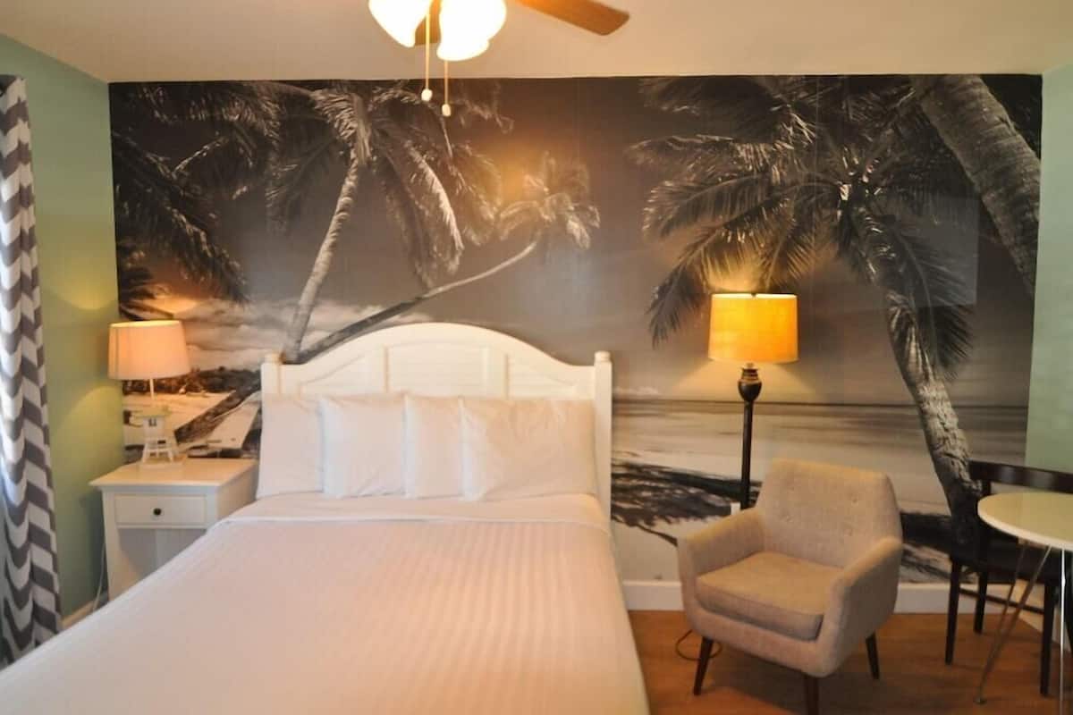 A hotel room with a bed, a chair, a table, and a wall mural of palm trees.