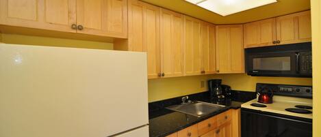 A compact kitchen with wooden cabinets, a white refrigerator, a black microwave, a stove, and a sink.
