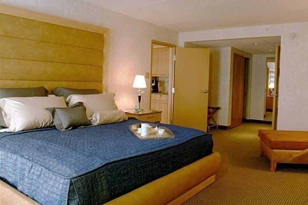 A hotel room with a large bed, a bedside table with a lamp, a small table with a tray, and a wooden bench.