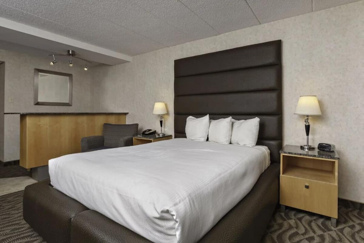 A hotel room with a large bed, two bedside tables with lamps, a wooden headboard, a grey armchair, and a mirror on the wall.