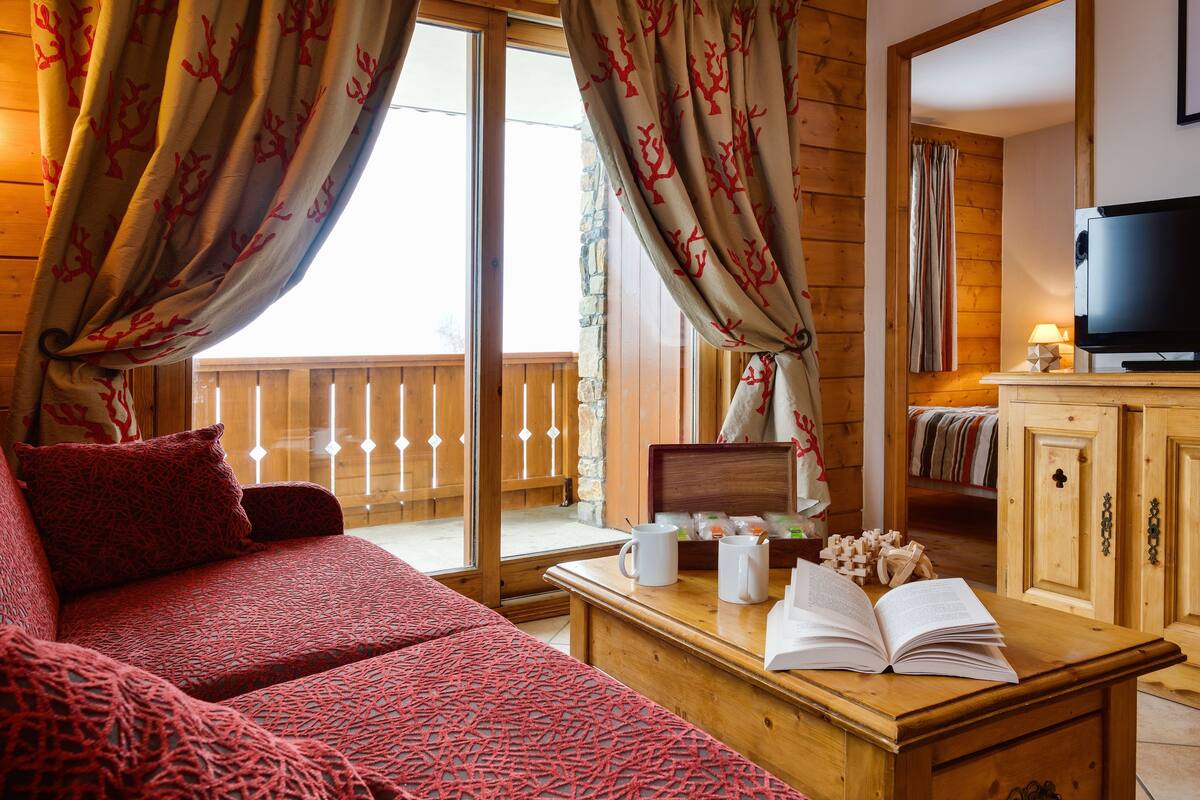 Ein gemütliches Zimmer mit einem roten, gemusterten Sofa, einem Holztisch mit einem Buch und Tassen, einem Fernseher und einem Balkon mit Holzgeländern.