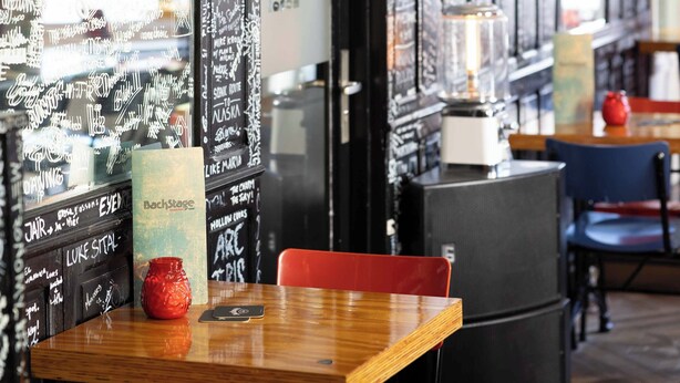 A restaurant interior with a saxophone mounted on the wall, a wooden table with a red vase, and a chalkboard menu. - Backstage Hotel Amsterdam (Amsterdam)