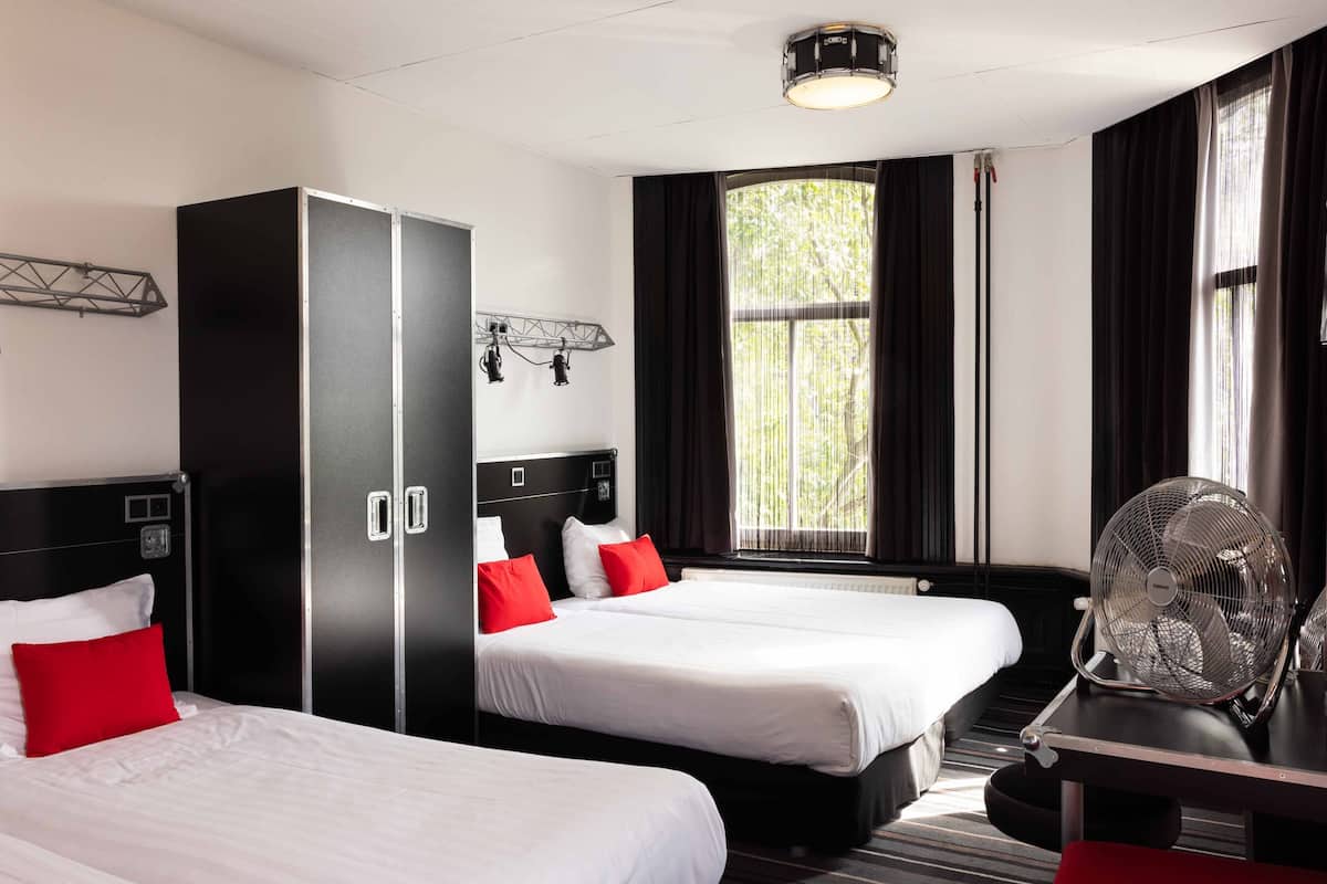 Ein Hotelzimmer mit zwei Betten, einem Kleiderschrank, einem Schreibtisch mit Ventilator und einem Fenster mit Vorhängen.