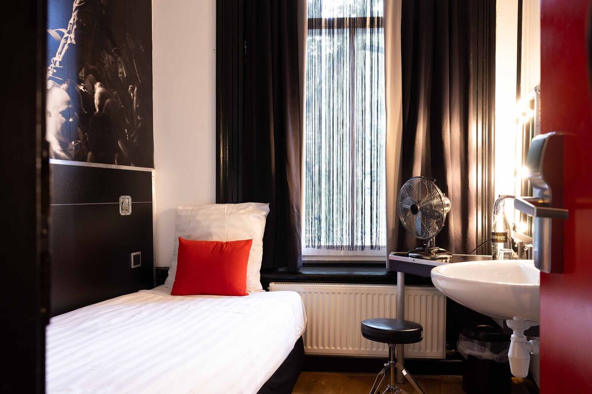 Ein Hotelzimmer mit einem Bett, einem roten Kopfkissen, einem Fenster mit Vorhängen, einer Heizung, einem Ventilator und einem Waschbecken.