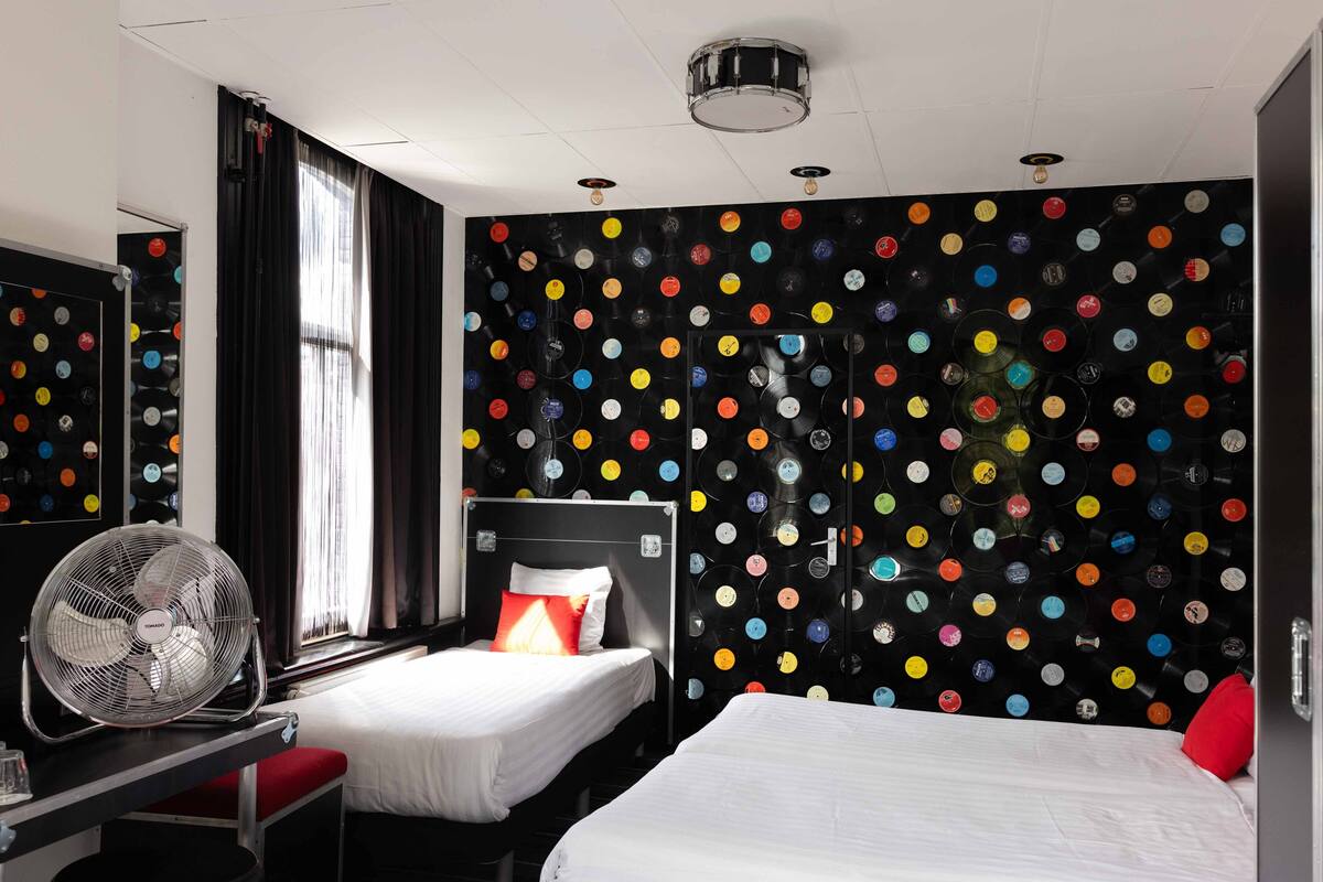 Ein Hotelzimmer mit zwei Betten, einem Schreibtisch mit Ventilator, einem Fenster mit Vorhängen und einer Wand, die mit bunten Flaschenverschlüssen verziert ist.