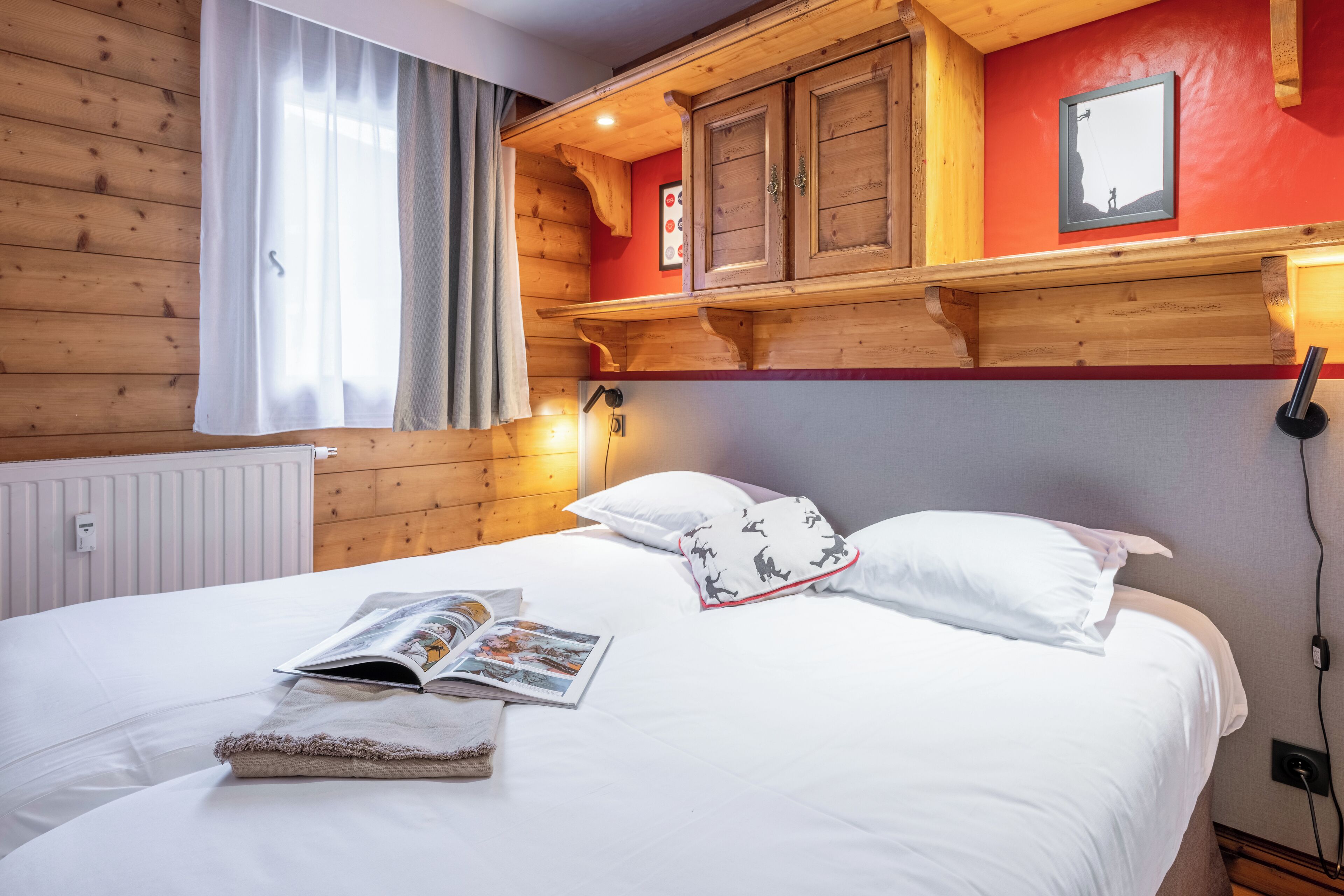 A neatly made bed with a magazine, a pillow with a pattern, and a wooden headboard with a shelf above.