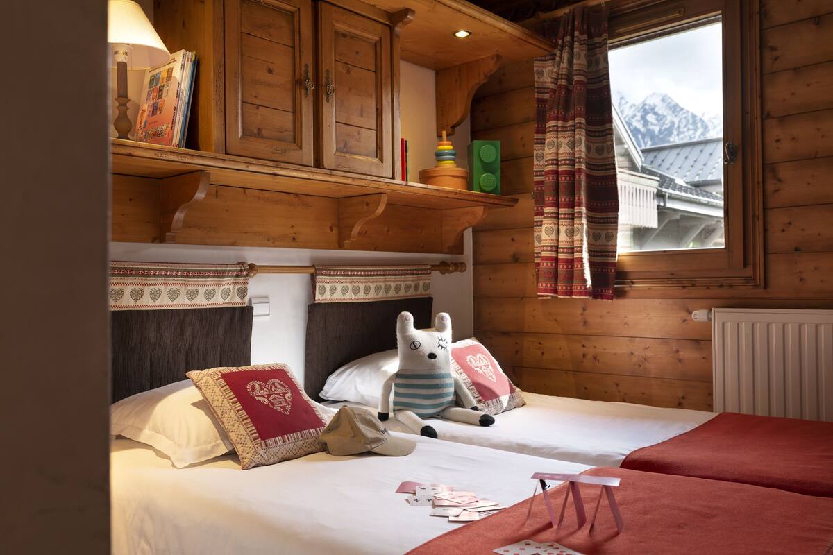 Ein Schlafzimmer einer Holzhütte mit zwei Betten, einer Lampe, Büchern und einem Fenster mit Blick auf Berge.