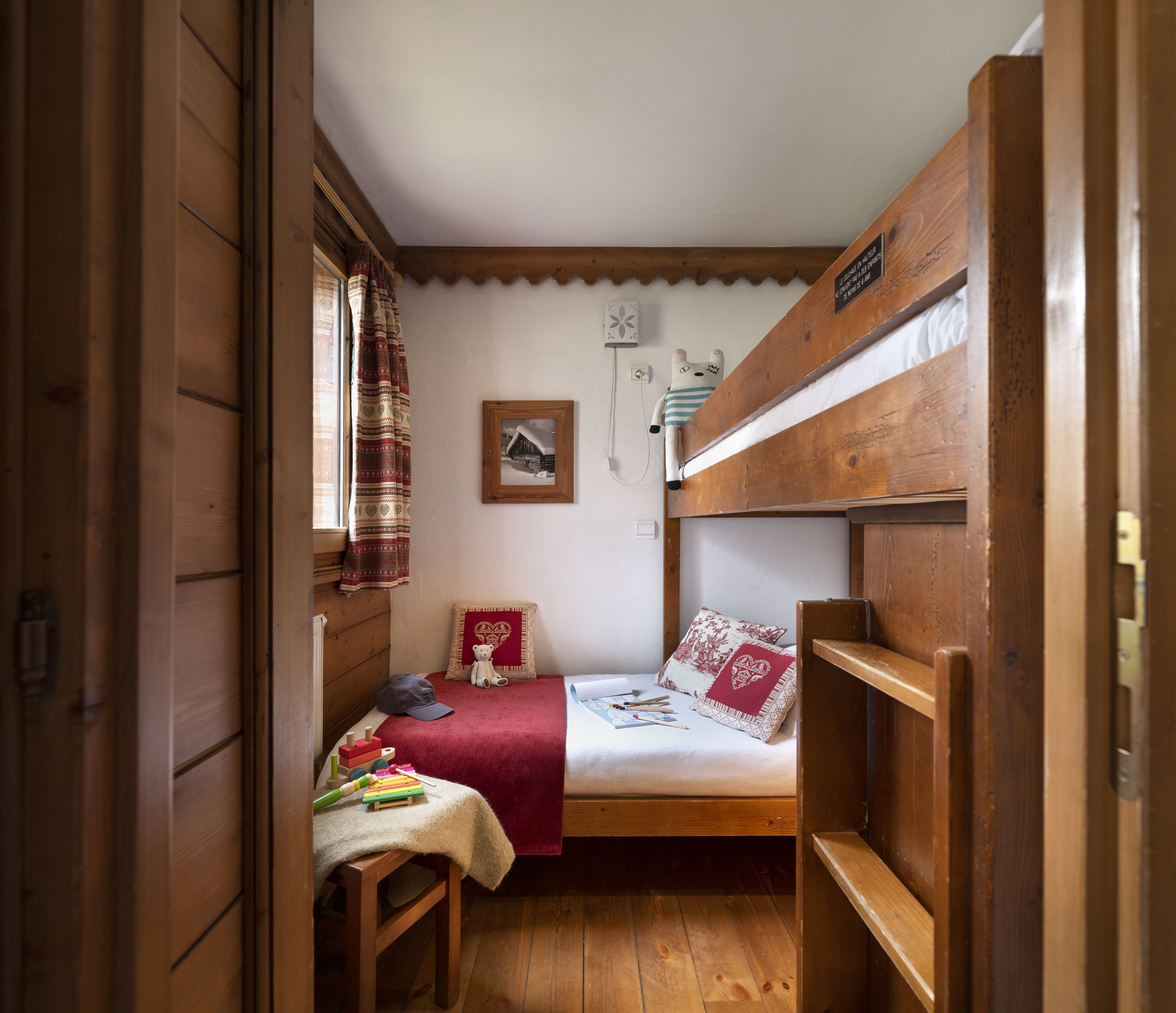 A cozy room with a bunk bed, a small desk with a chair, a window with curtains, and a wall-mounted light switch.