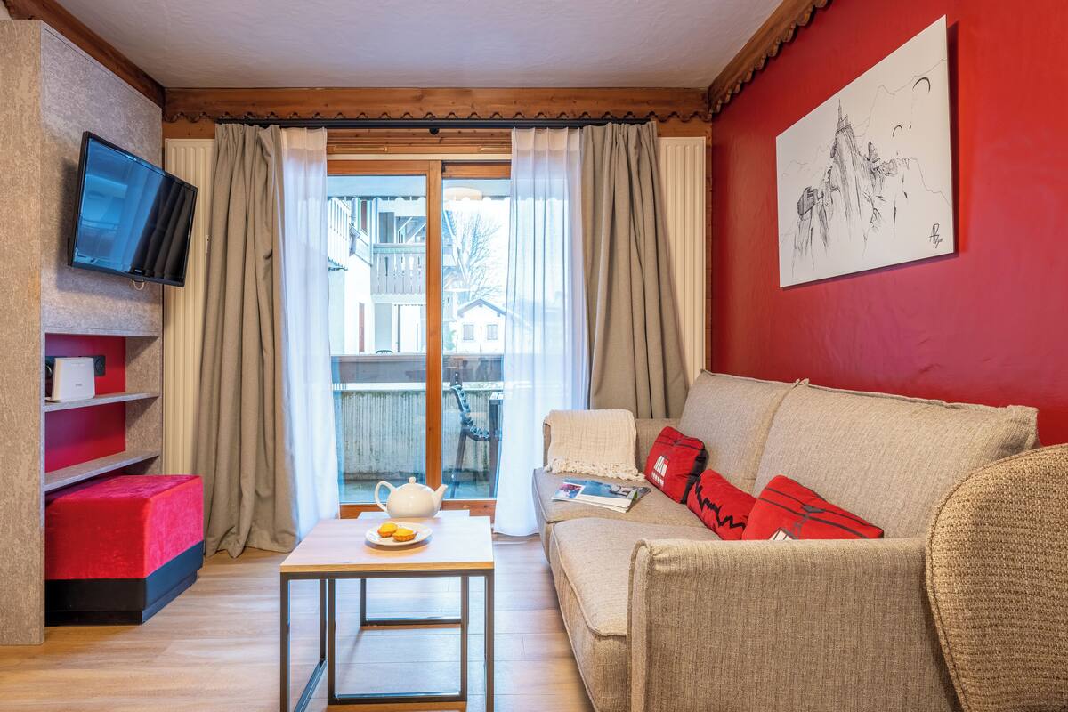 Ein gemütliches Wohnzimmer mit roter Wand, einem beigen Sofa, einem roten Hocker, einem kleinen Tisch mit Teekanne und einem an der Wand befestigten Fernseher.