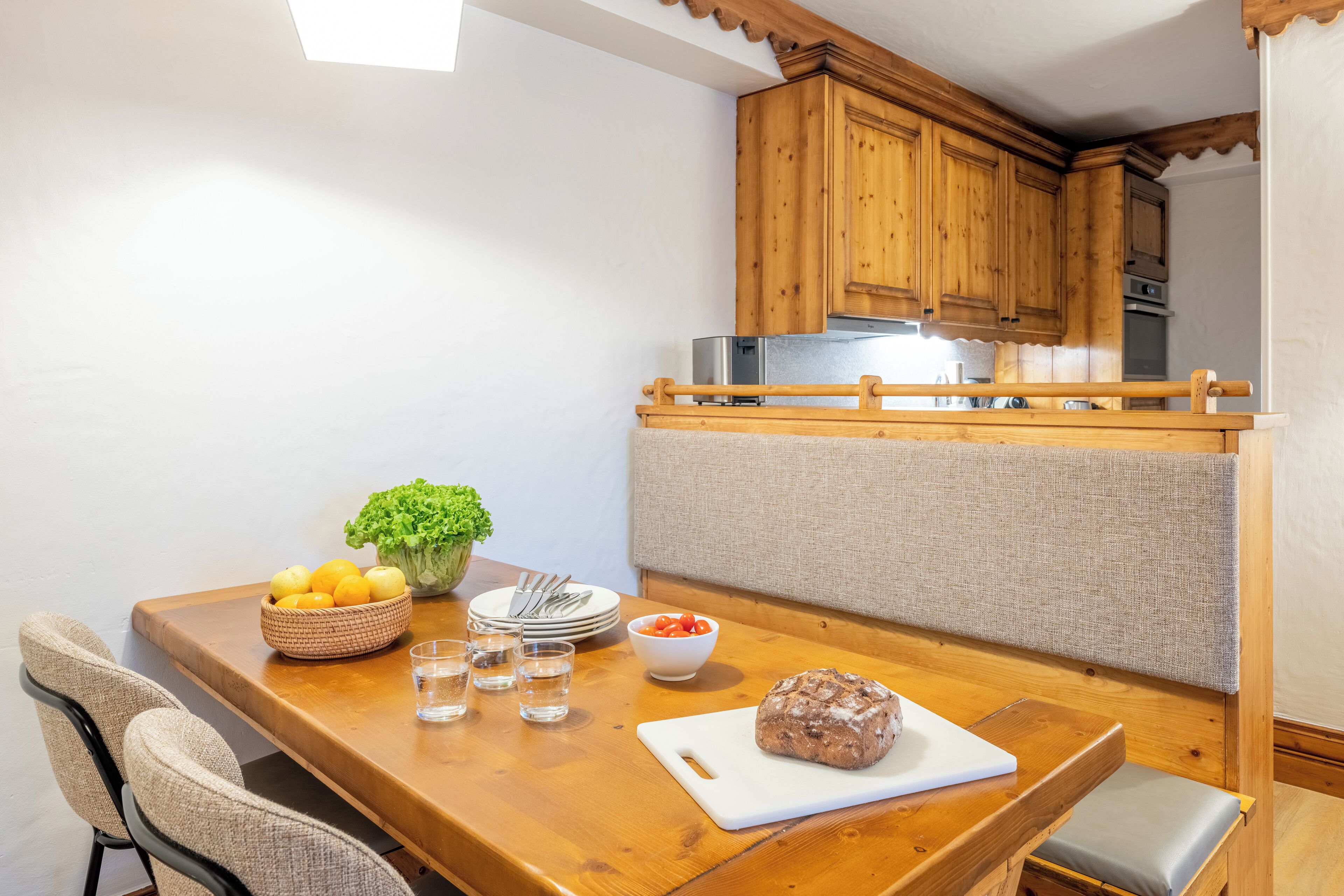 Een houten eettafel met een wit snijplank, een brood, een schaal met citroenen, een schaal met tomaten en een kleine plant.