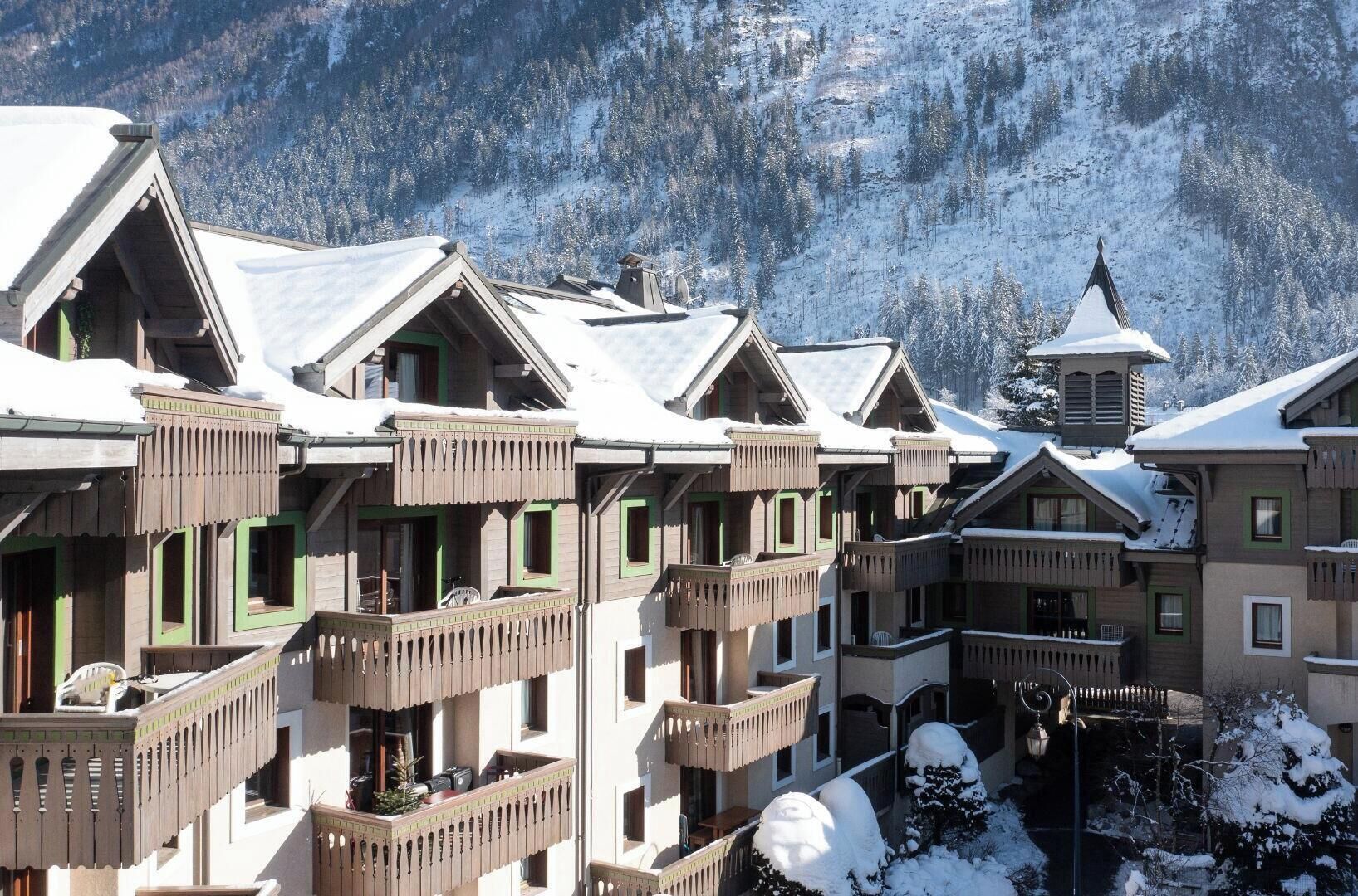 A row of chalet-style buildings with balconies, set against a snowy mountain backdrop.