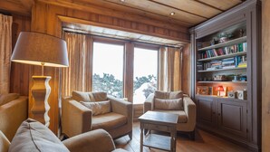 Un salón acogedor con paneles de madera, una estantería y vistas a árboles a través de amplias ventanas.