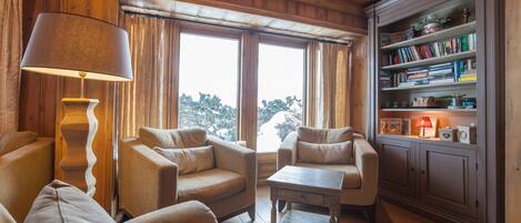 A cozy living room with wooden paneled walls, a bookshelf, and a view of trees through large windows.
