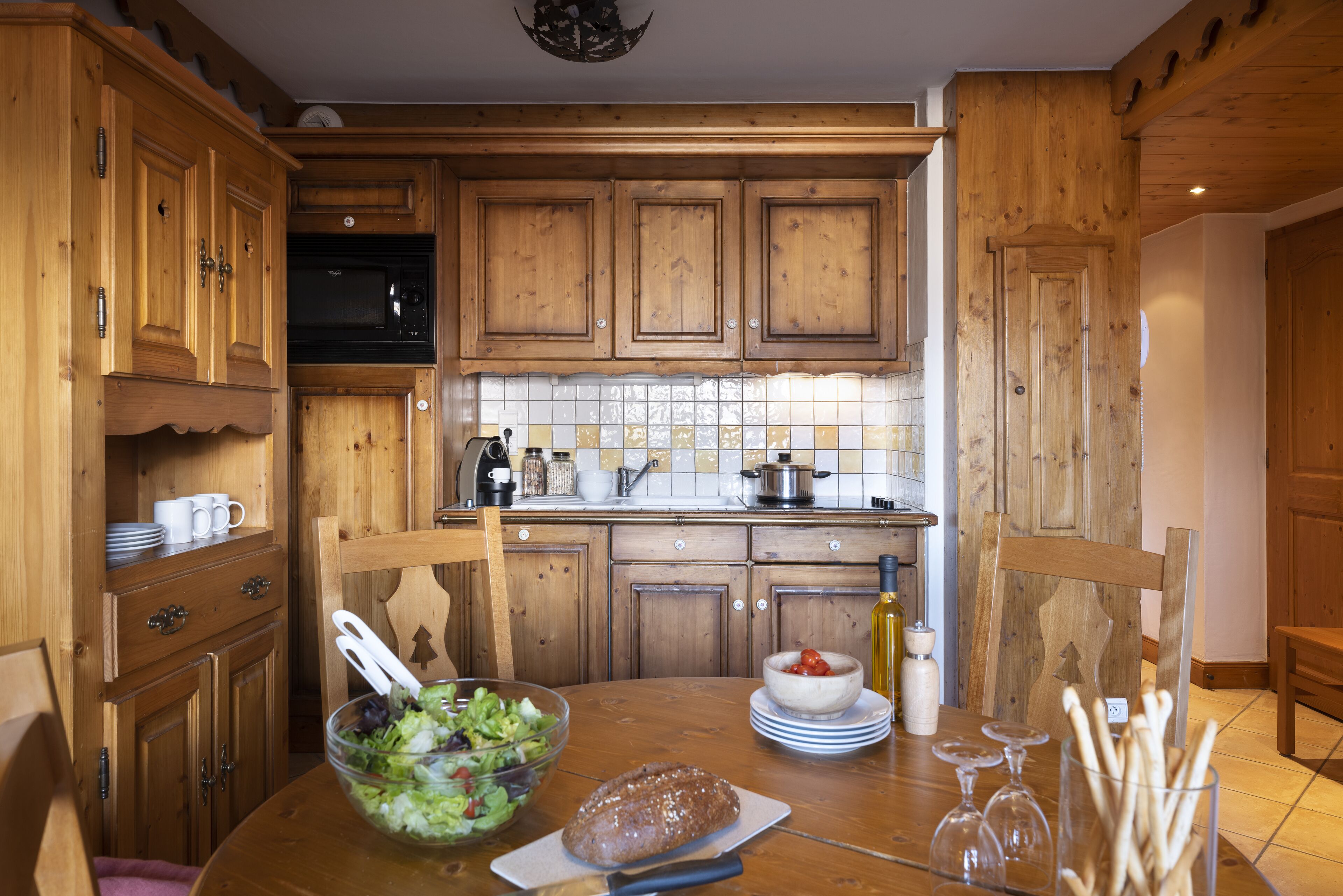 A wooden kitchen with cabinets, a dining table with a loaf of bread, a bowl of salad, and wine glasses.