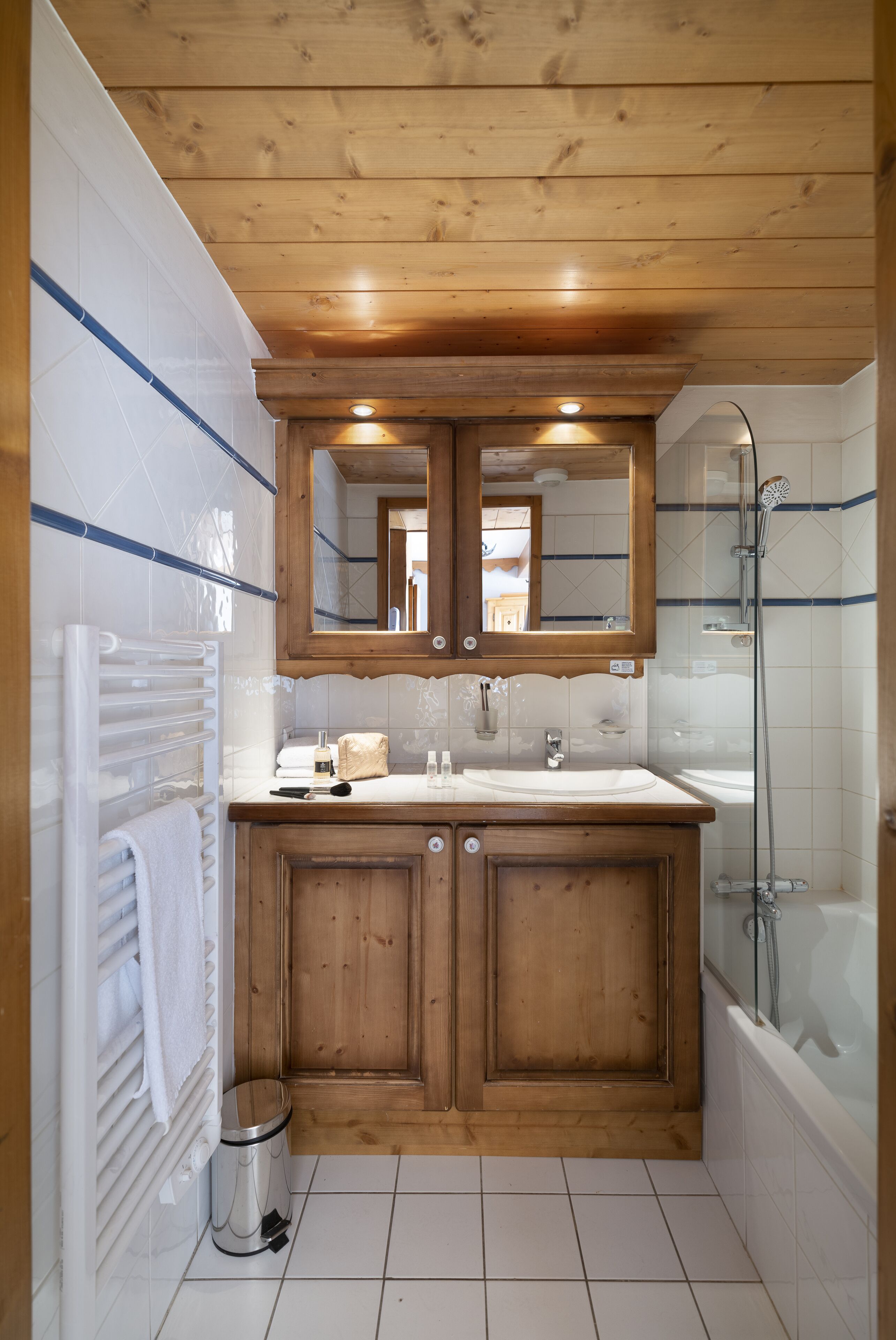 A wooden cabin bathroom with a double sink, a mirror above the sink, a shower, and a white tiled floor.