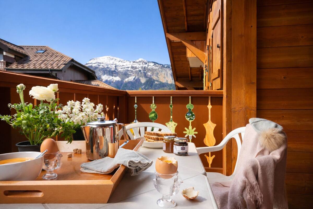 A wooden deck with a table set for breakfast, a view of snow-capped mountains, and a wooden cabin structure.