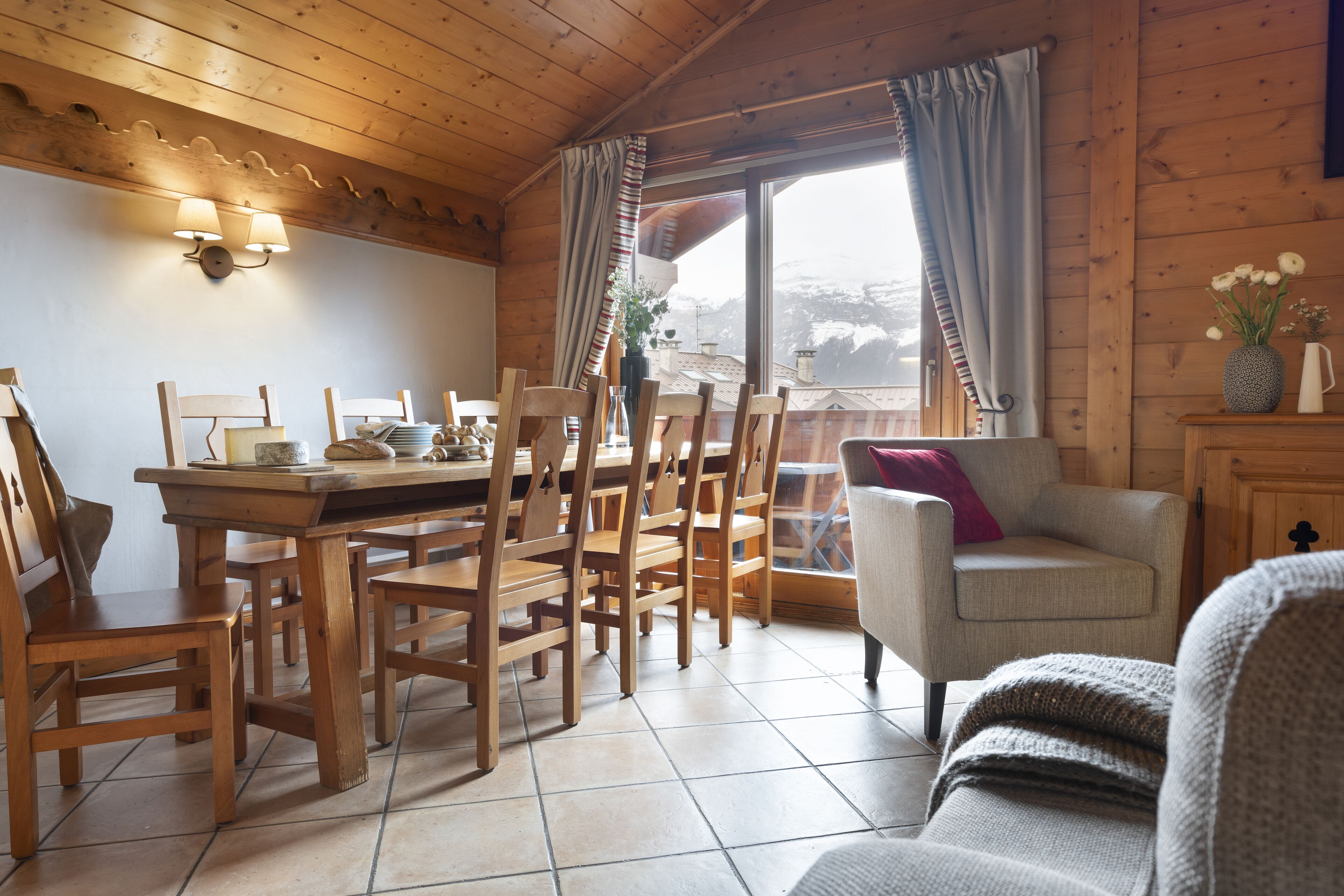 A wooden interior with a dining table and chairs, a sofa, and a window with curtains.
