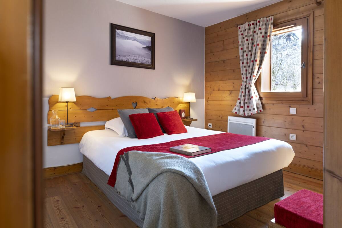 A wooden-paneled bedroom with a bed, two bedside lamps, a window with curtains, and a framed picture on the wall.