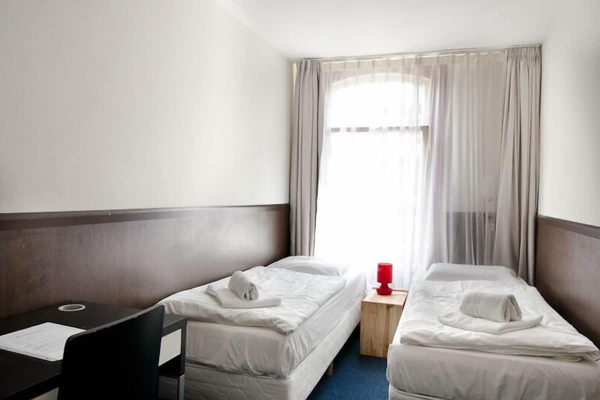 Habitación de hotel con dos camas individuales, un escritorio pequeño y una ventana con cortinas traslúcidas.