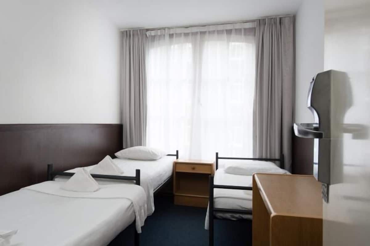 Habitación de hotel con dos camas individuales, cabecero de madera, un escritorio pequeño de madera, una silla y un ventanal con cortinas ligeras.