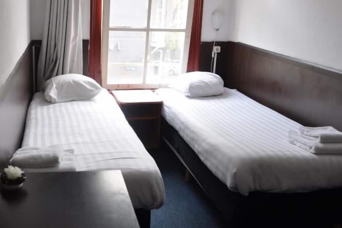 Una habitación de hotel pequeña con dos camas individuales, una ventana con cortinas y una mesita con un jarrón.