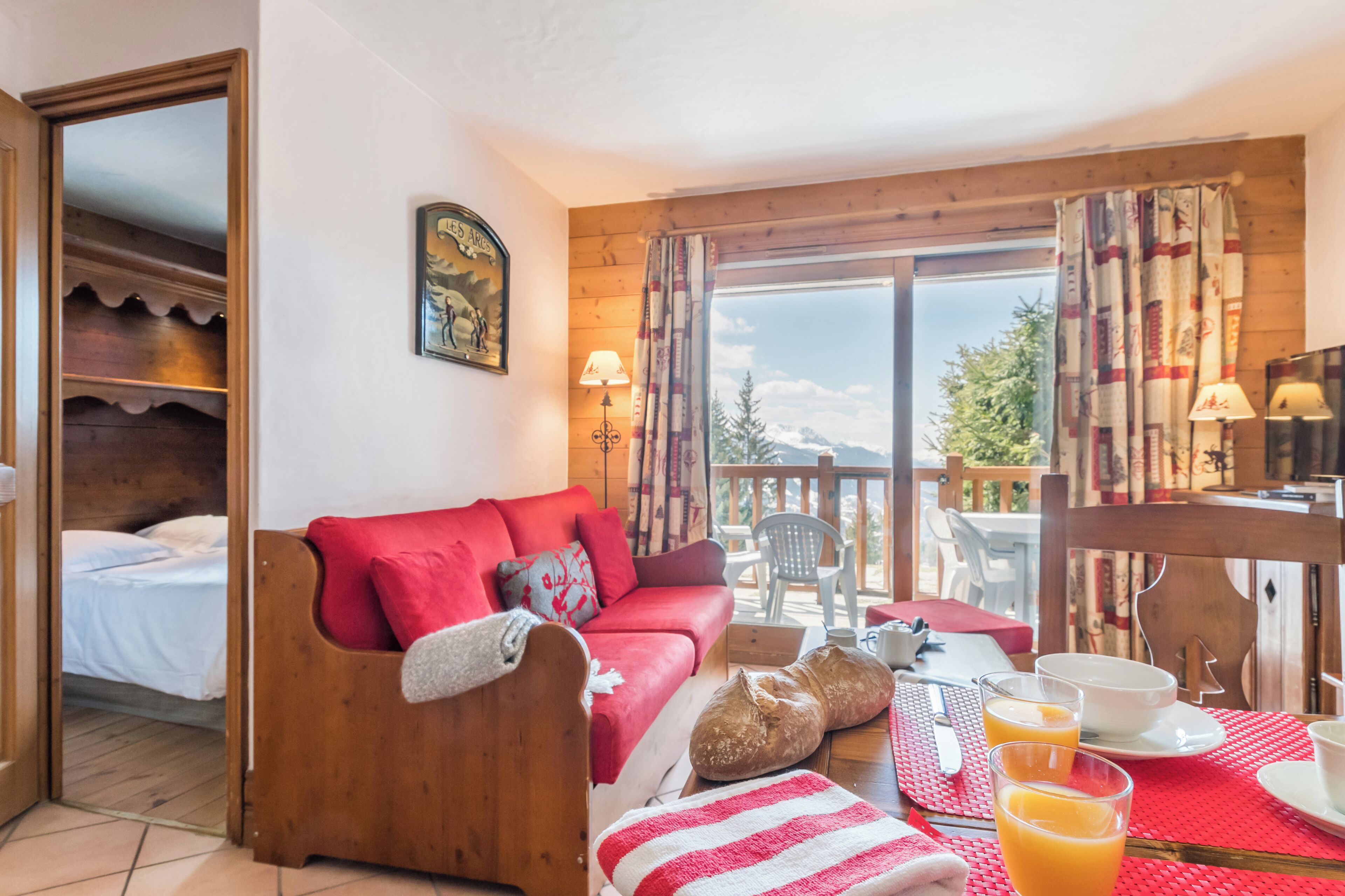 A cozy room with a red sofa, a wooden bed, a dining table with breakfast items, and a view of the outdoors.