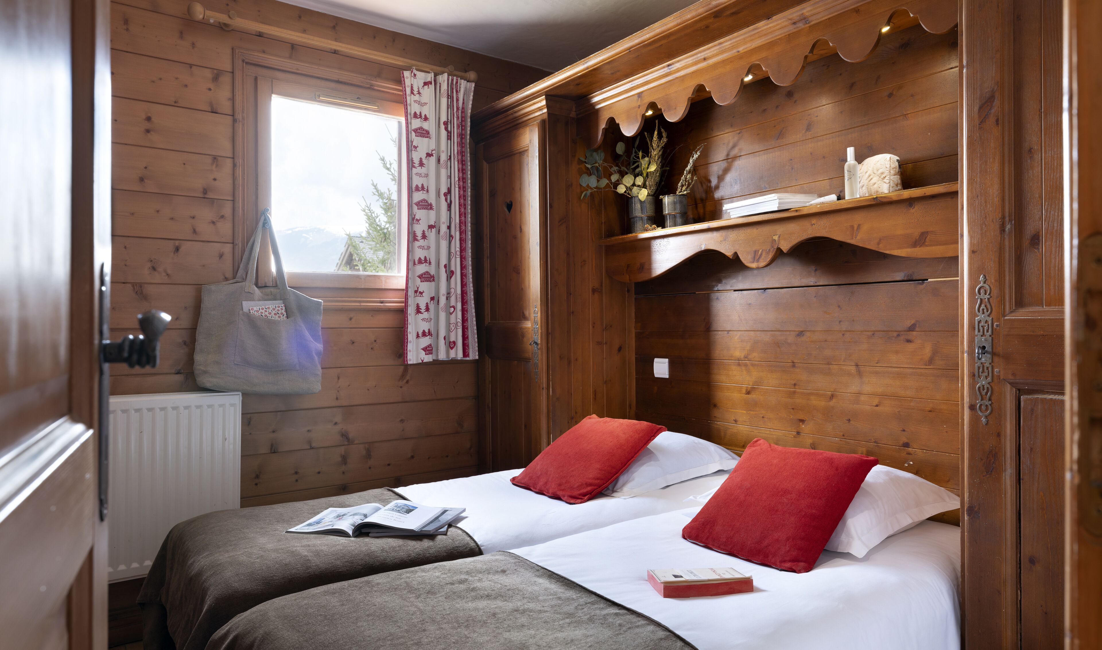 A wooden room with a bed, a window with curtains, and a shelf with decorative items.