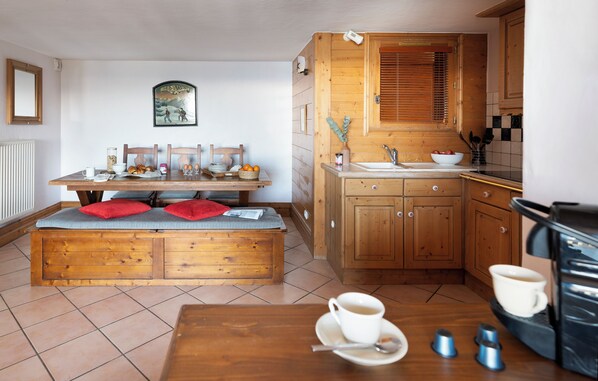 A cozy kitchen with wooden cabinets, a dining table with chairs, and a bench. There is a coffee machine and cups on the table.