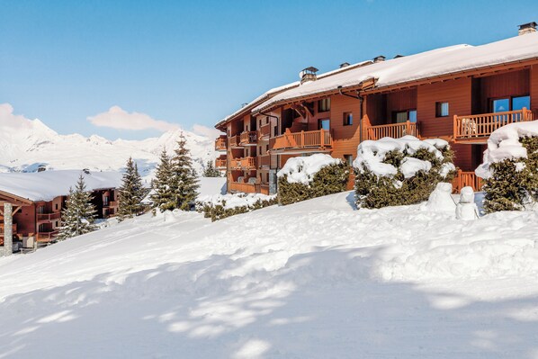 A multi-story wooden building with balconies, surrounded by snow-covered trees and mountains. - Résidence Pierre & Vacances Premium Les Alpages de Chantel (Bourg-Saint-Maurice)