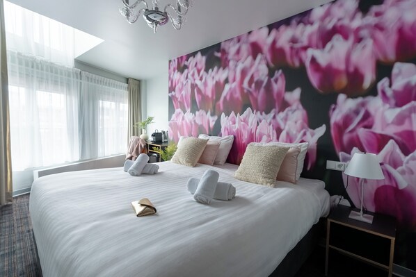 A hotel room with a large bed, white bedding, a wall mural of pink flowers, a bedside lamp, and a window with sheer curtains. - Hotel Multatuli (Amsterdam)