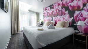 Habitació d'hotel amb un llit gran, televisió penjada a la paret, un penjallum i una paret amb un mural floral.