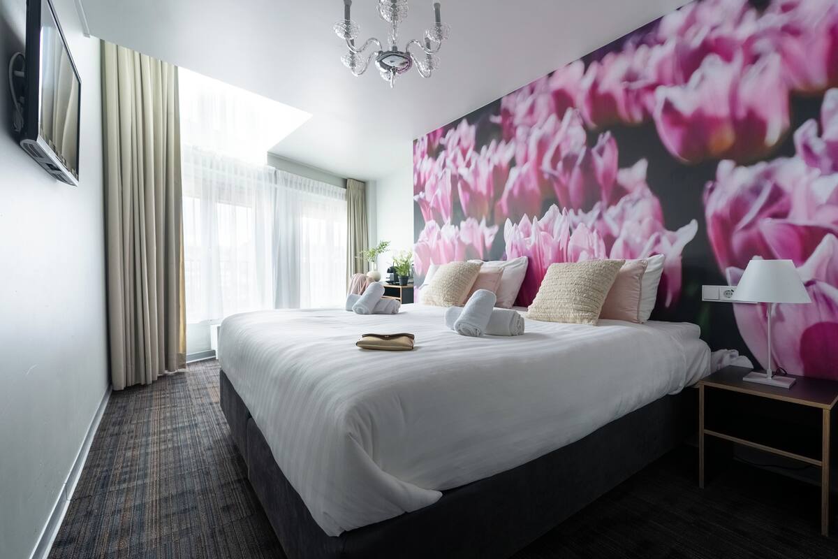 Habitació d'hotel amb un llit gran, televisió penjada a la paret, un penjallum i una paret amb un mural floral.