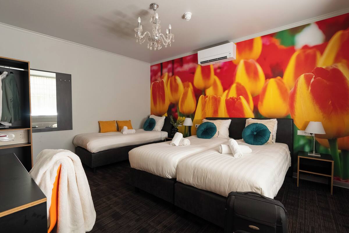 Habitació d'hotel amb dos llits, escriptori, cadira, armari, un lampadari i aire condicionat, i un mural floral de tulipes.