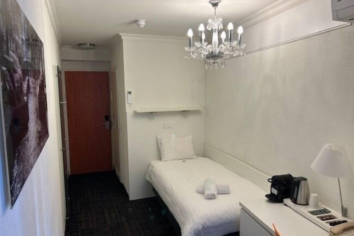 Habitació d'hotel amb un llit, una taula de nit amb una cafetera, una làmpada i un penjallum.